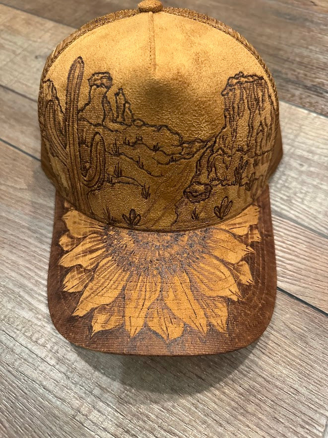 Brown cap with engraved desert scene and sunflower design on a wooden surface